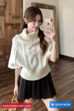 Woman taking a mirror selfie wearing a white cable knit sweater and black skirt, with a wooden floor and wall in the background.
