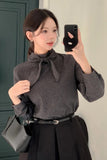 Woman taking a mirror selfie wearing a patterned blouse with a bow and black skirt.