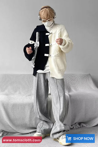 Person wearing a stylish outfit with a white sweater, gray pants, and black jacket in a minimalistic room.