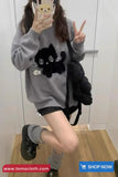 Person wearing a gray sweater with a black cat design, holding a phone with a Rilakkuma case, and posing in a room.