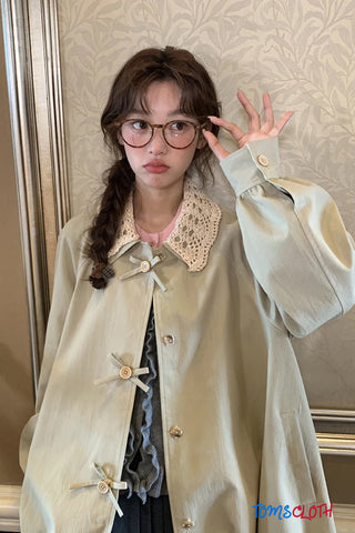 Person wearing a light-colored coat with decorative buttons and collar, standing against a patterned wall.