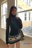Woman wearing a dark sweater with a patterned skirt standing in front of a window.