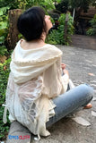 Person wearing a light-colored shawl sitting on a stone path with greenery around.
