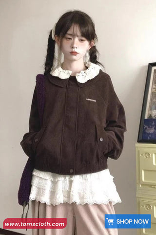 Woman wearing a brown jacket with a white collar and skirt, standing against a plain wall.