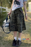 Person wearing a camouflage skirt, white t-shirt with graphic design, and black sneakers, holding a bag outdoors.