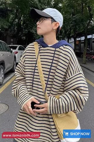 Person wearing a striped sweater and cap, holding a phone, with a blurred background of a street scene.