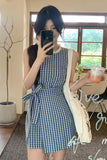 Woman in a blue checkered dress taking a selfie in a room with a window and plant.