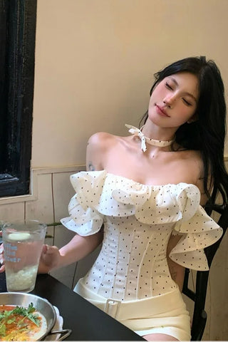 Woman in a white off-shoulder dress sitting at a table with a drink and food.