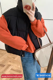 Person wearing an orange hoodie and black puffer vest holding a phone, with a beige handbag on a wooden floor.