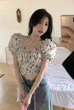Woman taking a mirror selfie wearing a floral top and denim skirt in a bedroom.