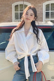 Woman in a white blouse and blue jeans standing next to a car with a brick building in the background