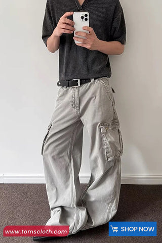 Person wearing light gray cargo pants and a dark shirt, holding a phone, against a white wall.