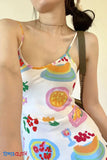 Woman wearing a colorful fruit-patterned dress with 'TOMS CLOTH' branding.