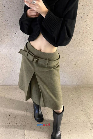 Person wearing a black sweater and green skirt with a belt, standing against a beige wall.