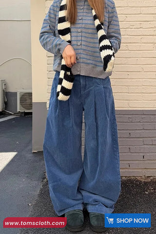 Person wearing a striped sweater, blue jeans, and holding a black and white striped scarf against a beige wall.