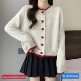 Person wearing a white cardigan with red heart buttons and a black skirt indoors.