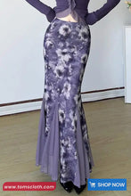 Person wearing a long, patterned purple dress against a white wall.