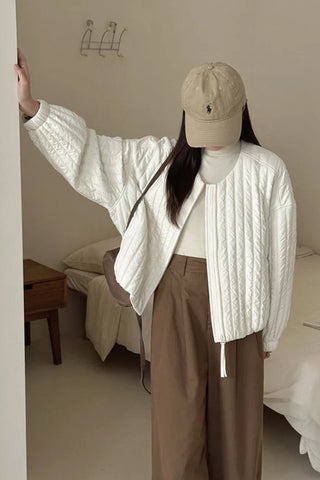 Person wearing a white cable knit sweater and beige cap in a room with neutral decor.