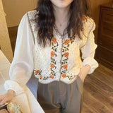 Person wearing a white blouse with an embroidered floral vest in a room.
