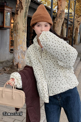 Person wearing a floral jacket, brown beanie, and holding a beige handbag in an outdoor setting.