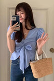 Woman wearing a blue top and jeans, holding a phone and a woven bag indoors.