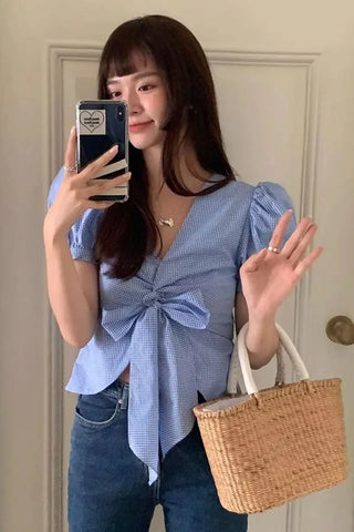 Woman wearing a blue top and jeans, holding a phone and a woven bag indoors.