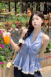Woman in a blue lace dress holding a drink outdoors with plants and a wooden structure in the background.