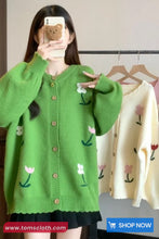 Person wearing a green cardigan with floral patterns, holding a phone with a bear case.