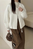 Person wearing a white textured jacket and brown pants holding a brown bag indoors.