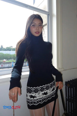 Woman wearing a black turtleneck and patterned skirt standing indoors by a window.