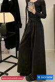 Person wearing a long black coat and wide-leg pants, standing in a room with a lamp and clothing rack.