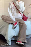 Person wearing a white cardigan, brown pants, and red sneakers sitting on a bed.
