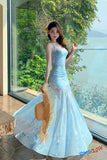Woman in a light blue dress holding a straw bag by a large window with greenery and water view.
