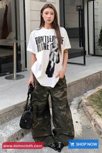 Woman wearing a white t-shirt with text and camouflage pants, standing on a sidewalk.