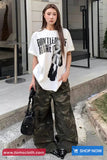 Woman wearing a white t-shirt with text and camouflage pants, standing on a sidewalk.