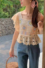 Woman wearing a crochet top and blue jeans, standing outdoors with greenery in the background.