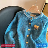Blue knitted cardigan with a heart-shaped brooch on a hanger against a neutral background.