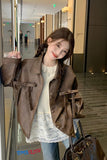 Woman in a brown coat standing in an indoor setting with a handbag.