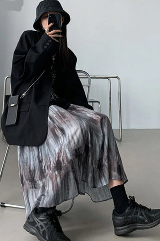 Person wearing a black coat, patterned skirt, and black shoes, sitting on a chair.
