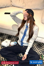 Person sitting on a couch with a phone and handbag, wearing a white sweater and black cap.