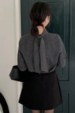 Person wearing a gray checkered blouse with a bow tie and black skirt, standing in front of a dark green door.