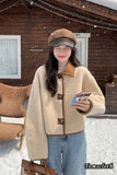 Person wearing a beige jacket with brown accents in front of a wooden building.