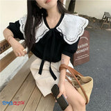 Woman sitting on a bench wearing a black and white outfit with a lace collar, holding a phone and a straw bag.