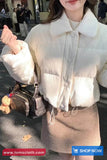 Person wearing a white textured jacket and beige skirt with a handbag, standing outdoors.