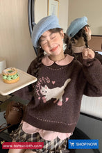 Child wearing a brown sweater with a cat design, blue beret, and plaid skirt, holding a donut.