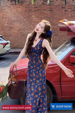 Woman in a blue floral dress standing next to a red car with a brick wall background.