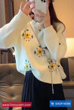 Person wearing a white sweater with floral patterns, holding a phone in an indoor setting.