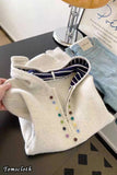 White jacket with colorful buttons held by a hand on a table with jeans and a lamp in the background.