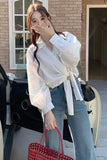 Woman wearing a white blouse and blue jeans, holding a red bag, standing in front of a brick building.