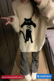 Person wearing a cream sweater with a black cat design, pointing at the sweater.